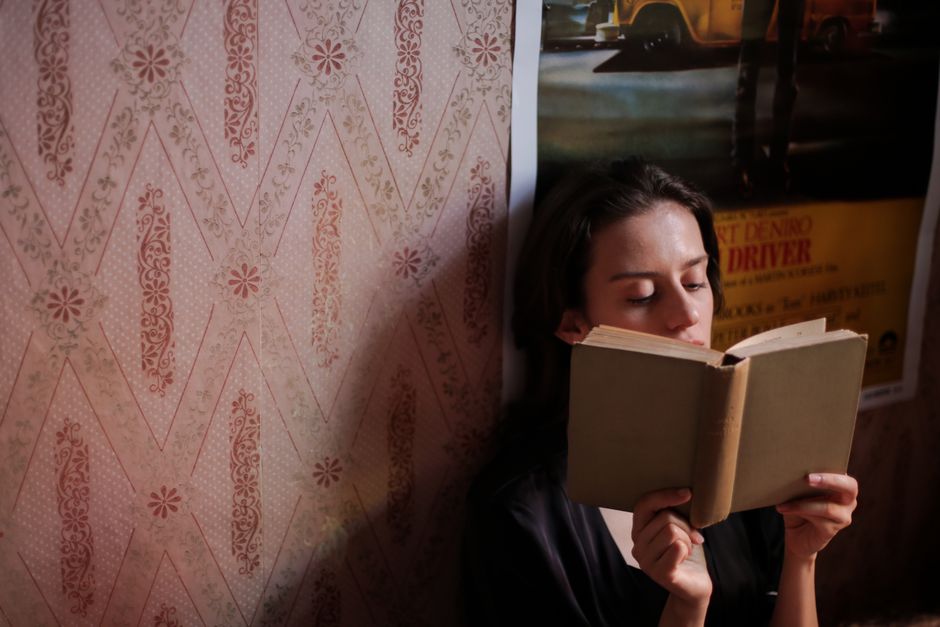A young woman deeply engrossed in a book, surrounded by vintage wallpaper indoors.