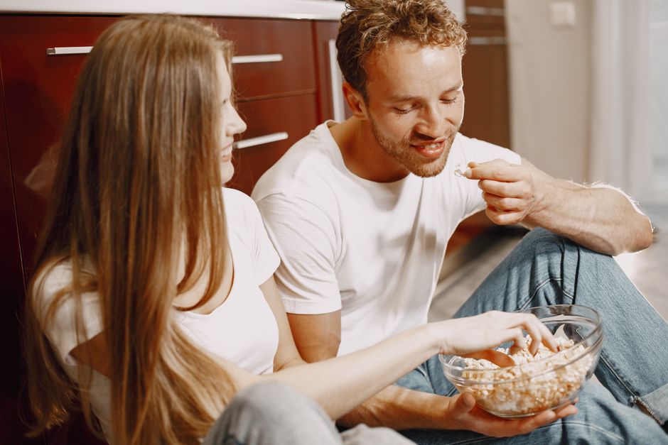 Happy couple sitting on the floor indoors, sharing a bowl of popcorn and smiling at each other.